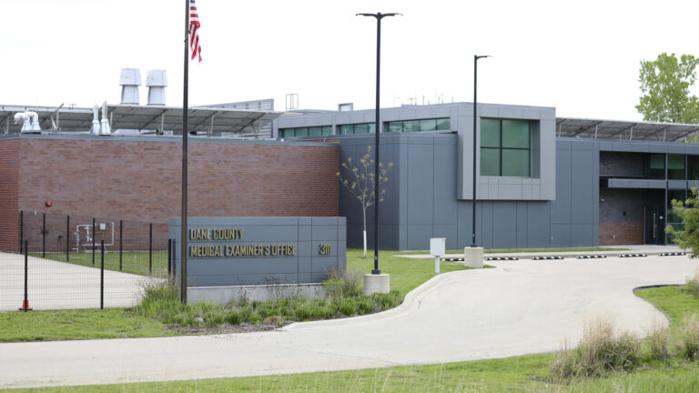 A two-story building with areas of brick wall, metal siding and few windows is fronted by a driveway and a sign reading Dane County Medical Examiner's Office.