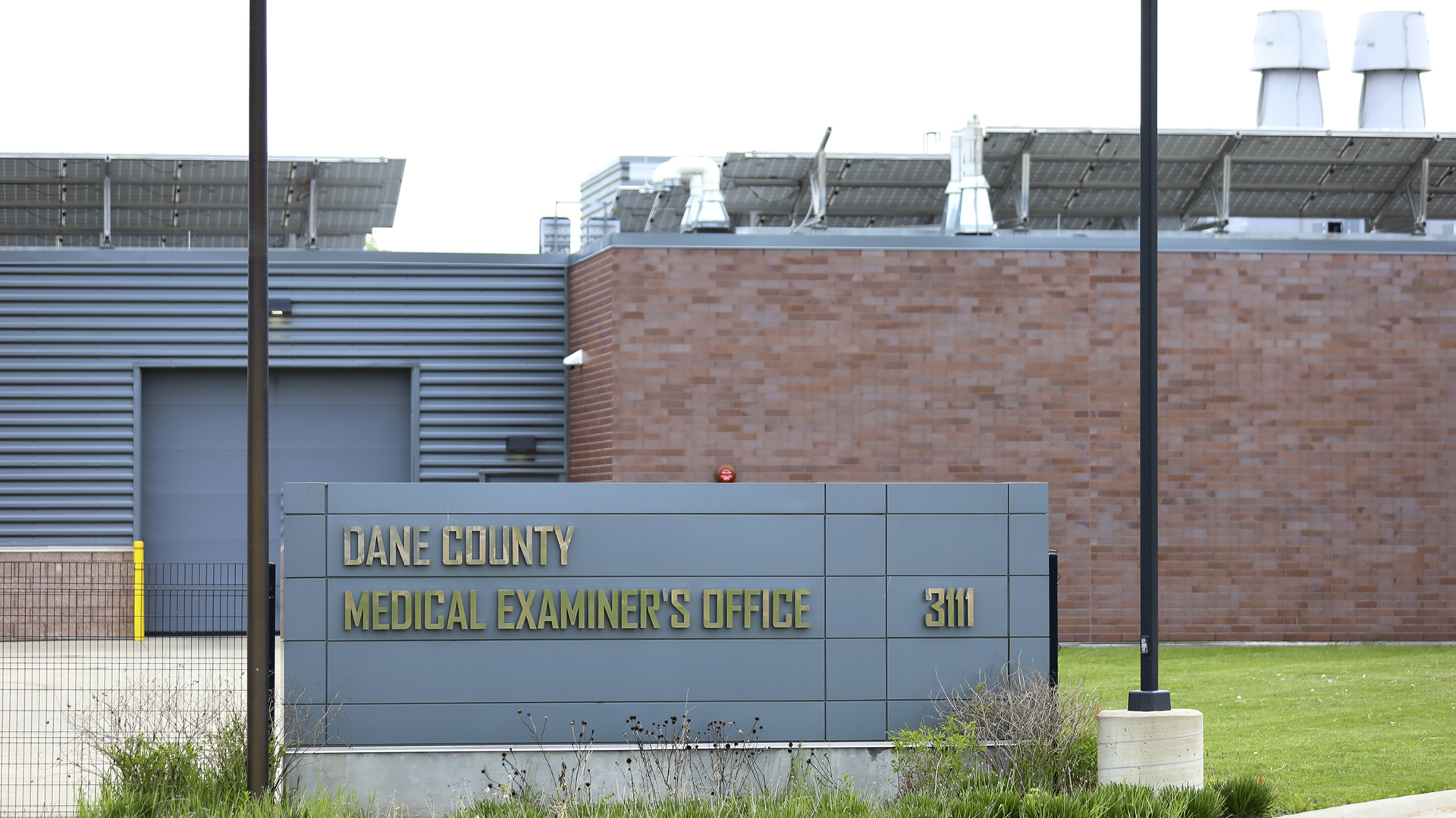 A two-story building with areas of brick wall, metal siding, few windows and rooftop solar panels is fronted by a driveway and a sign reading "Dane County Medical Examiner's Office."