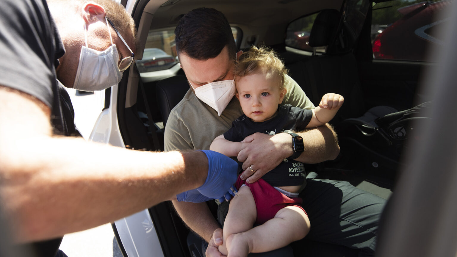 A toddler seated on the lap of an adult inside a car is given a shot in the thigh.