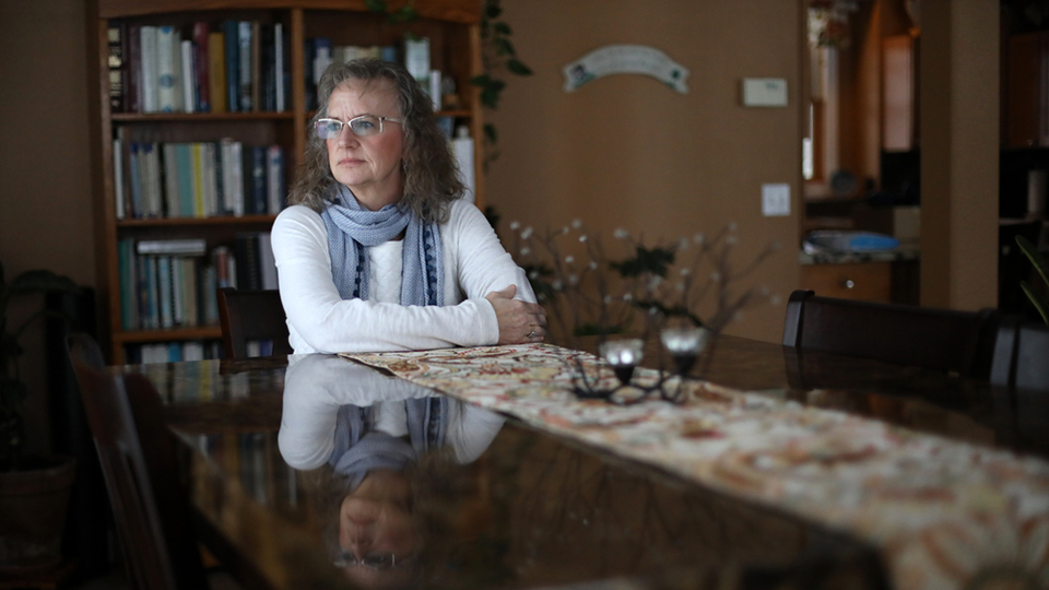 Alicia Cashman sits at a table in a dining room with her reflection on its polished surface and a bookshelf in the background.