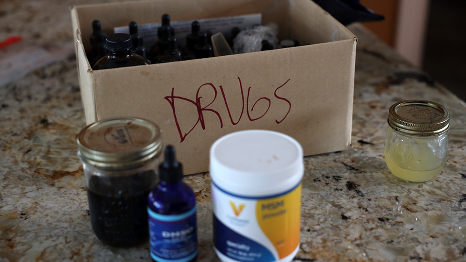 A box labeled "Drugs" and filled with vials and bottles stands on a countertop, with several other jars and containers placed outside of it.