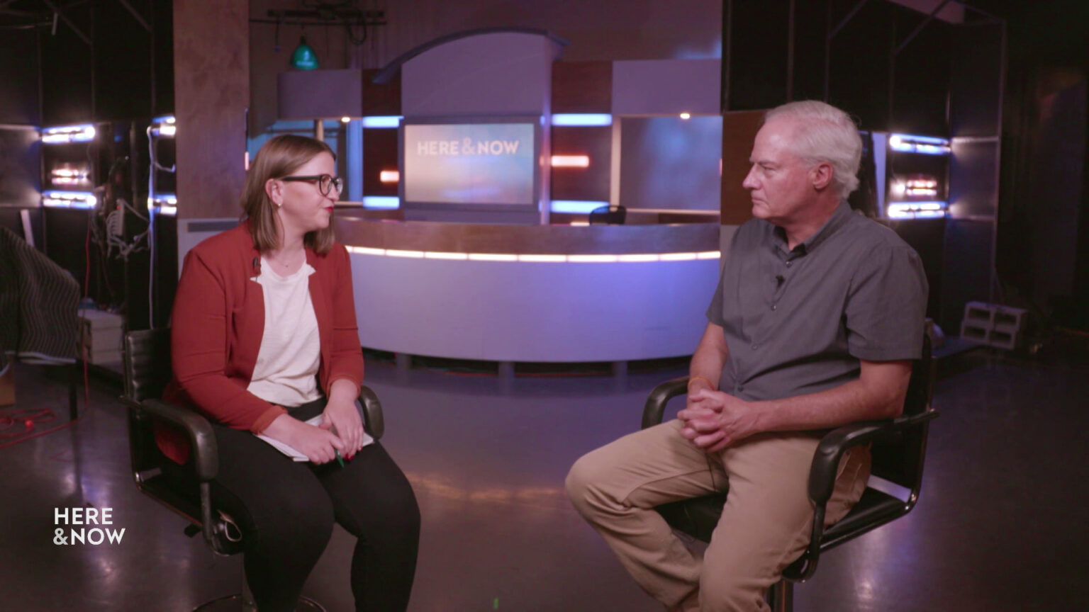 Marisa Wojcik and Dr. James Conway sit in chairs in a room with the Here & Now set in the background.