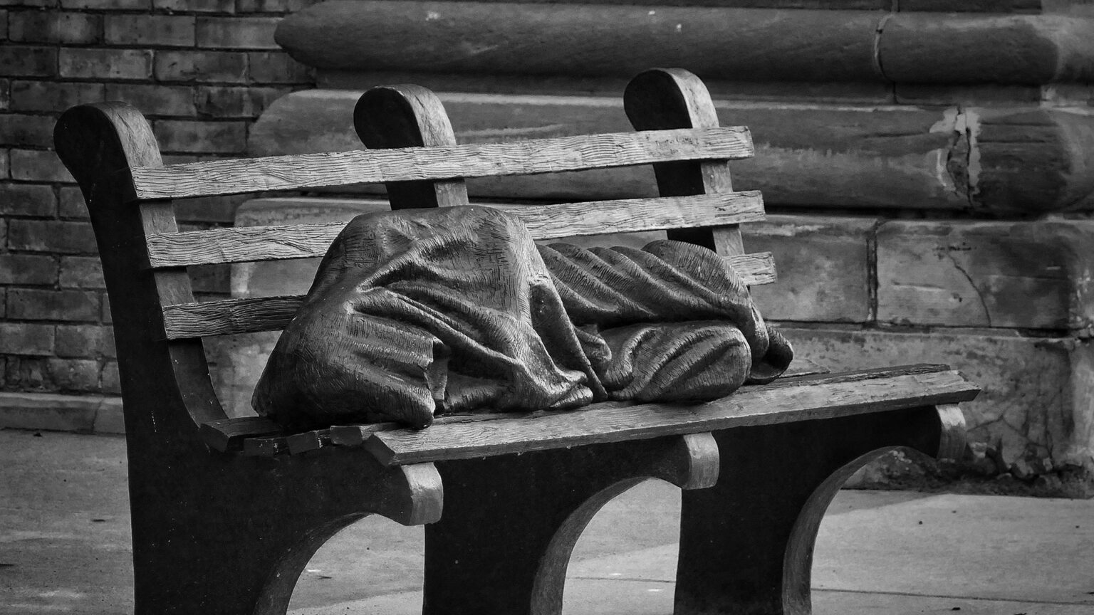 A sculpture depicts a sleeping person wrapped in a blanket laying on a wood park bench.