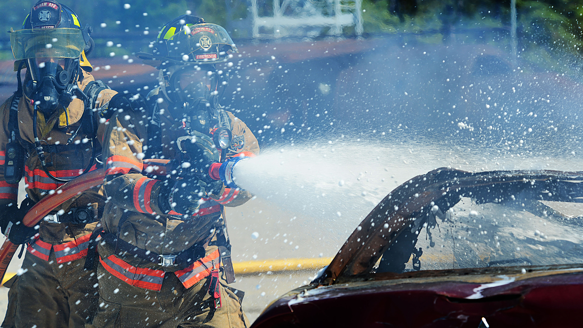 Two firefighters in full gear spray foam out of a hose onto a burned vehicle