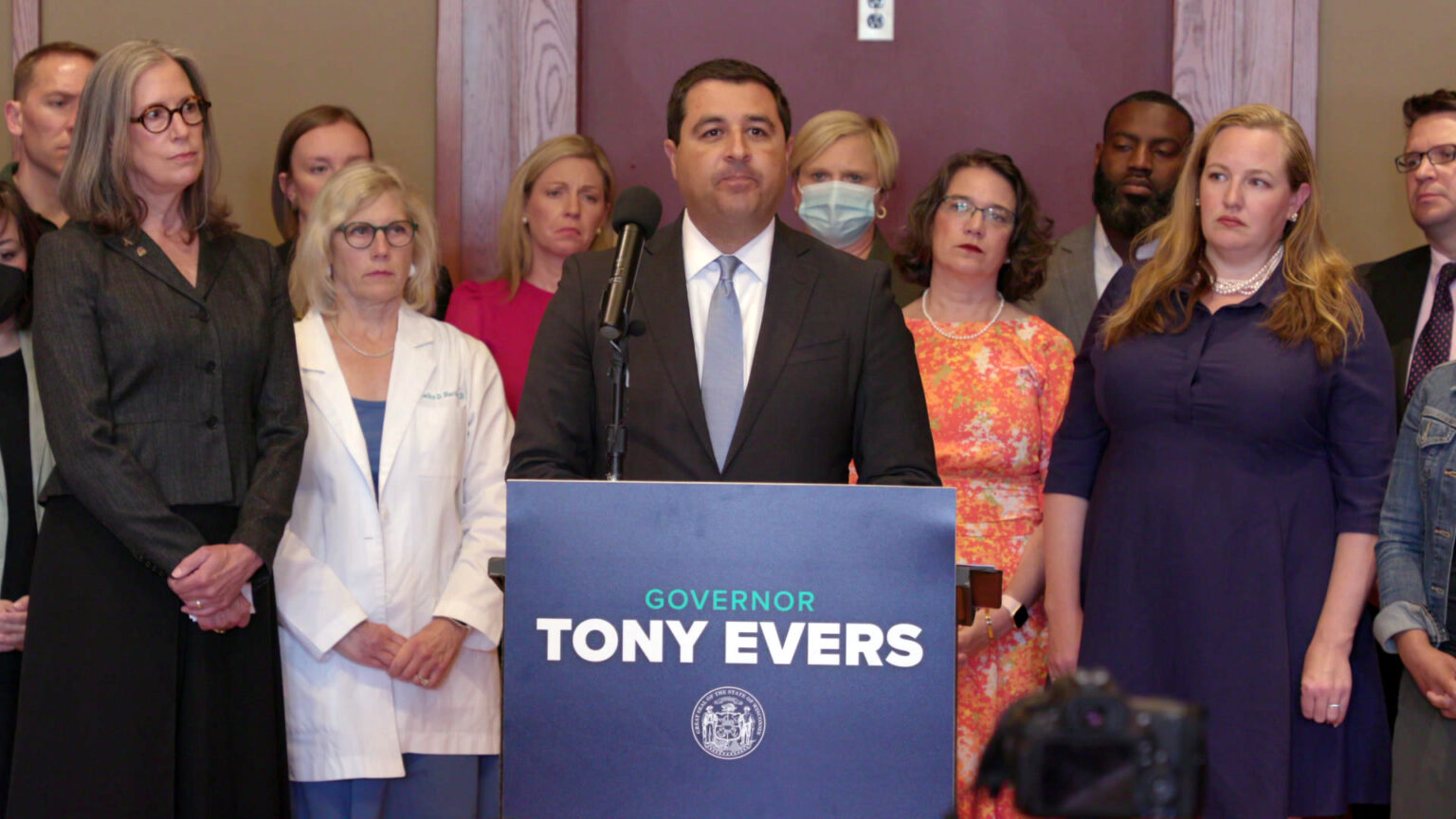Josh Kaul stands behind a podium with a sign reading Governor Tony Evers with a line of people standing in the background.