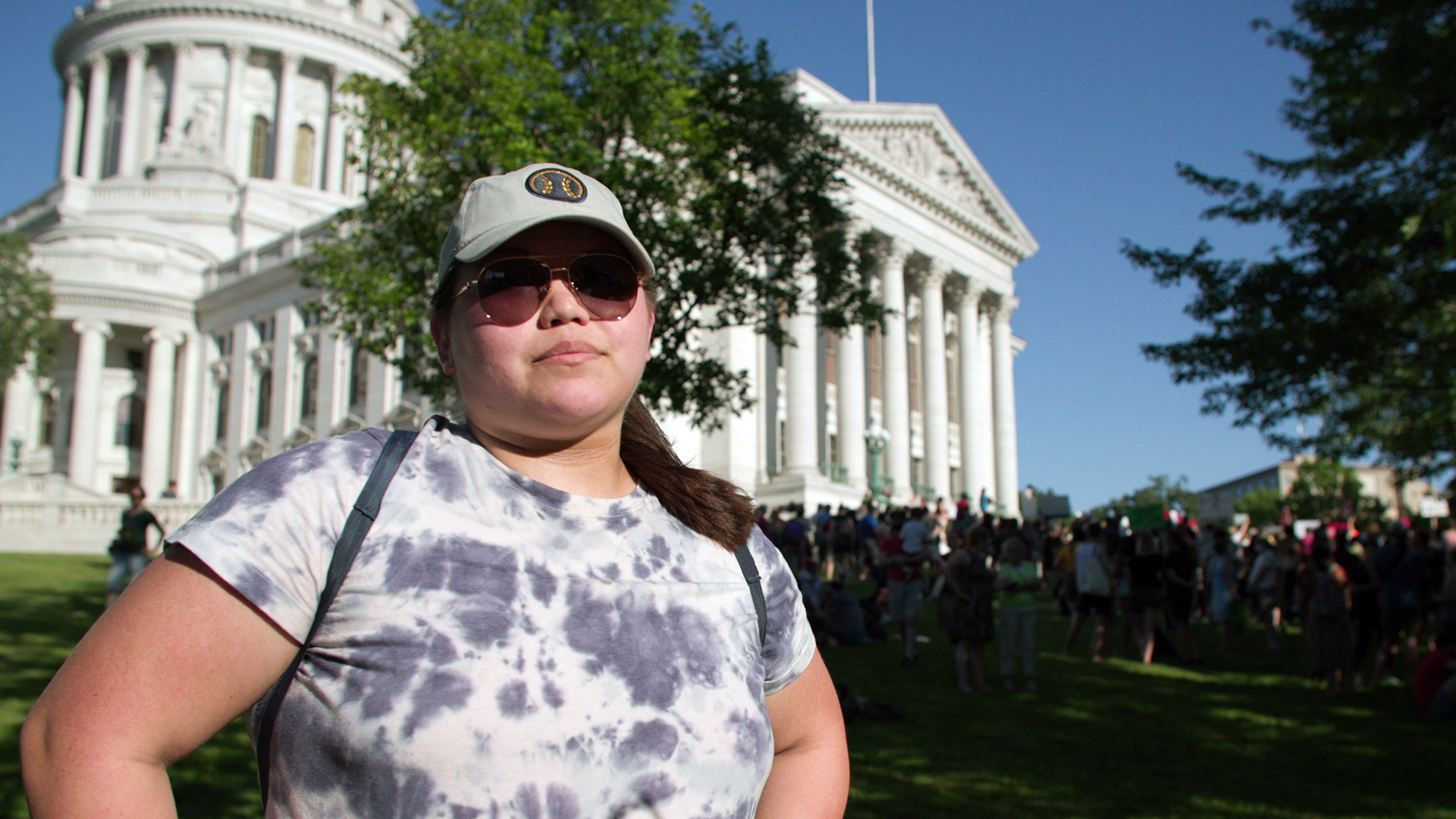 Alyssa Bhoopat stands in front of the Wisconsin State Capitol with protesters in the background.