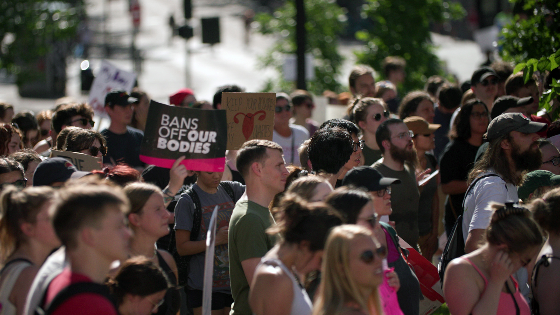 Protesters stand and listen to a speaker, with several holding signs.