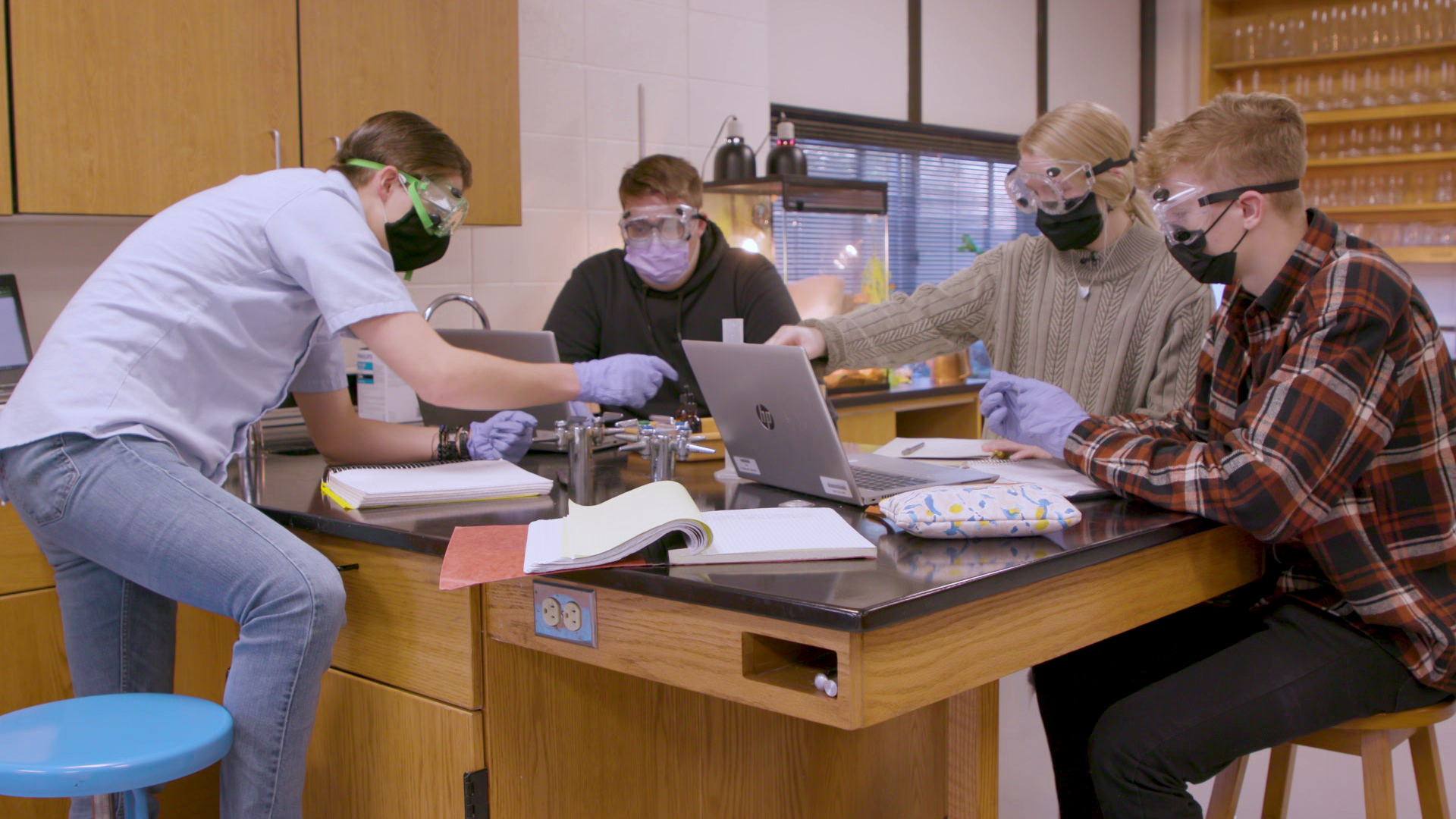 Students wearing safety goggles and face masks sit and stand around a science lab bench covered with laptop computers and books.