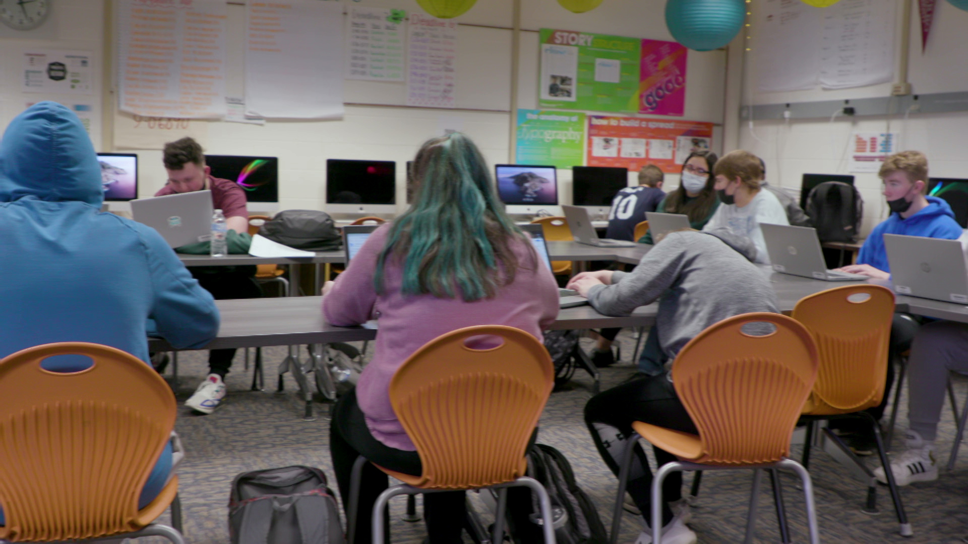 Students work on laptops while seated in chairs facing tables in a classroom.