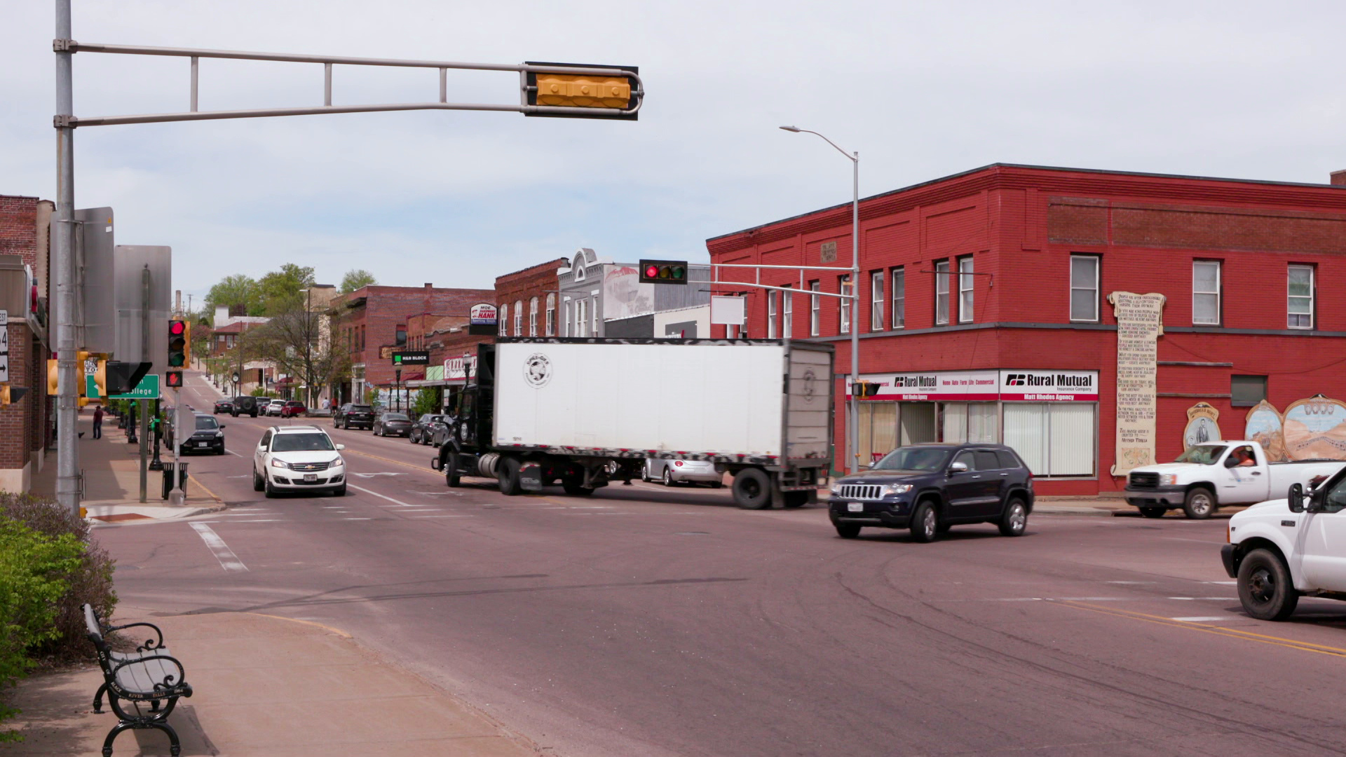 Personal vehicles and a cargo truck pass through an intersection with a stoplight in Black River Falls, surrounded by two-story buildings.