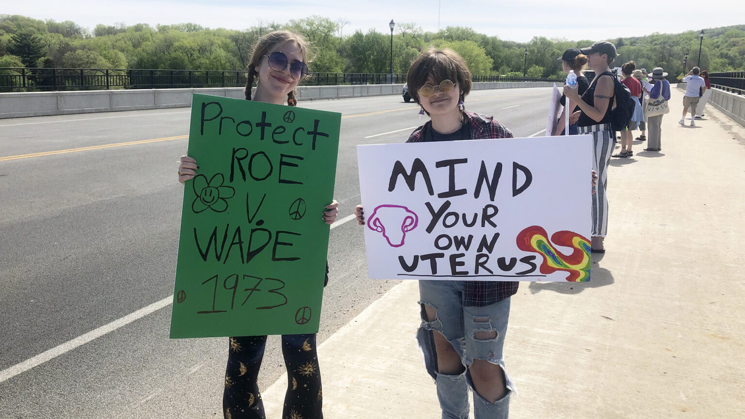 Two protestors standing on a bridge sidewalk hold signs reading Protect Roe v. Wade 1973 and Mind Your Own Uterus with other protestors standing in the background.