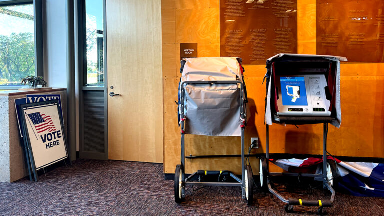 Two touchscreen printed ballot voting machines, one with its screen covered, stand in a room with wood-paneled walls and plaques listing the names of donors, with a U.S. flag on a poll on the floor and a stack of signs reading Vote Here leaning against a concrete wall under a plate glass window.