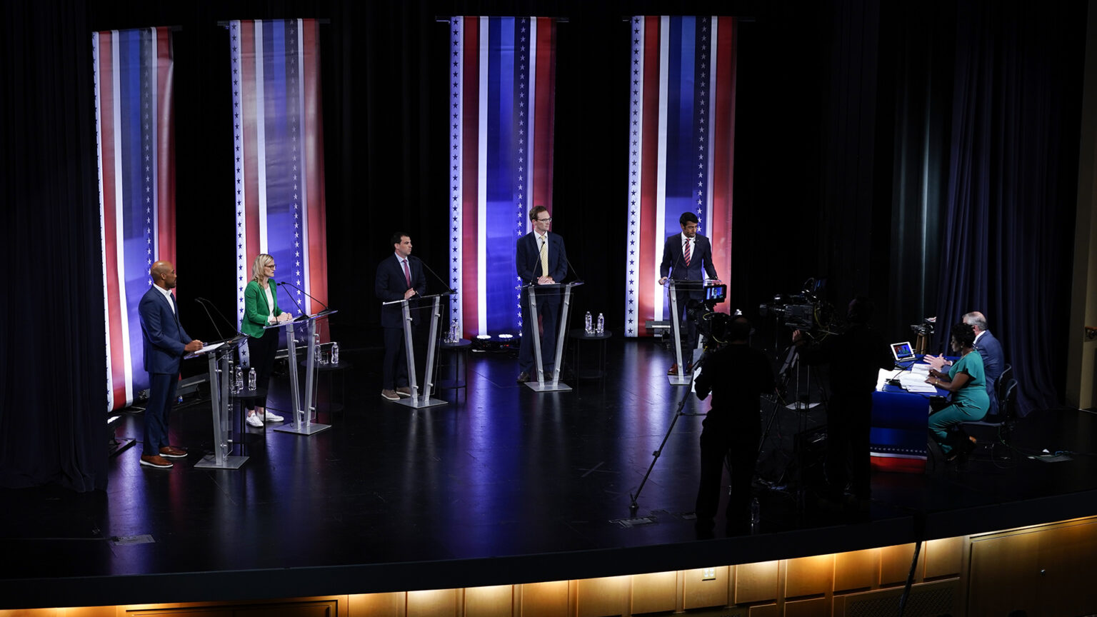 Mandela Barnes, Sarah Godlewski, Alex Lasry, Tom Nelson and Steven Olikara stand behind podiums on a stage facing television cameras and two seated moderators.