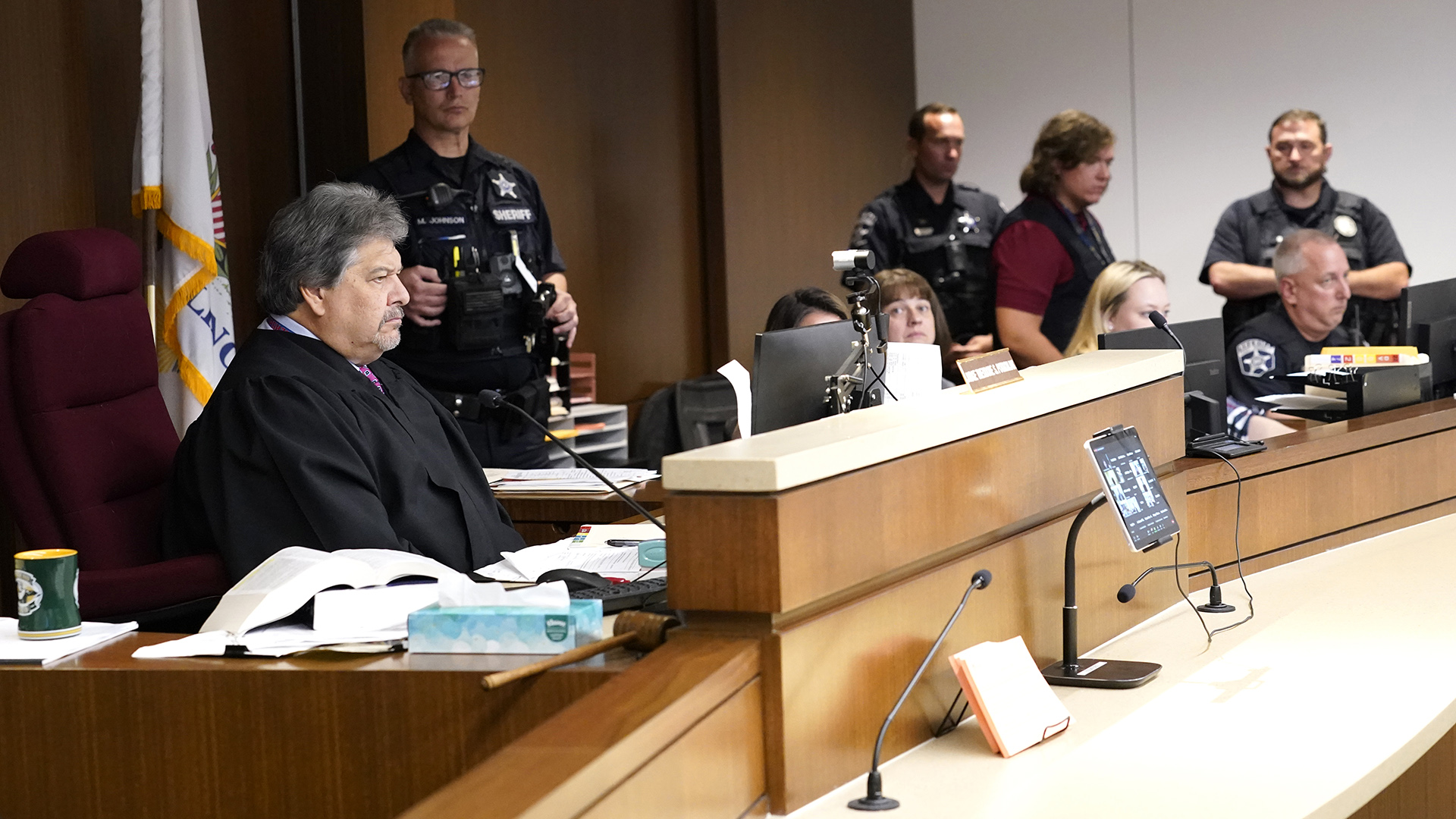 Judge Theodore Potkonjak sits behind a dais with courtroom officers and other staff in the background.