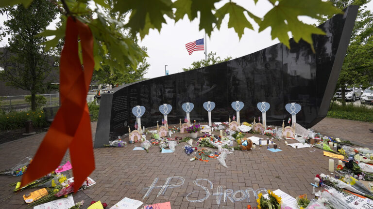 Flowers, posters and other items are placed on the ground in front of a large black stone sculpture, with a U.S. flag flying at half-staff in the background.