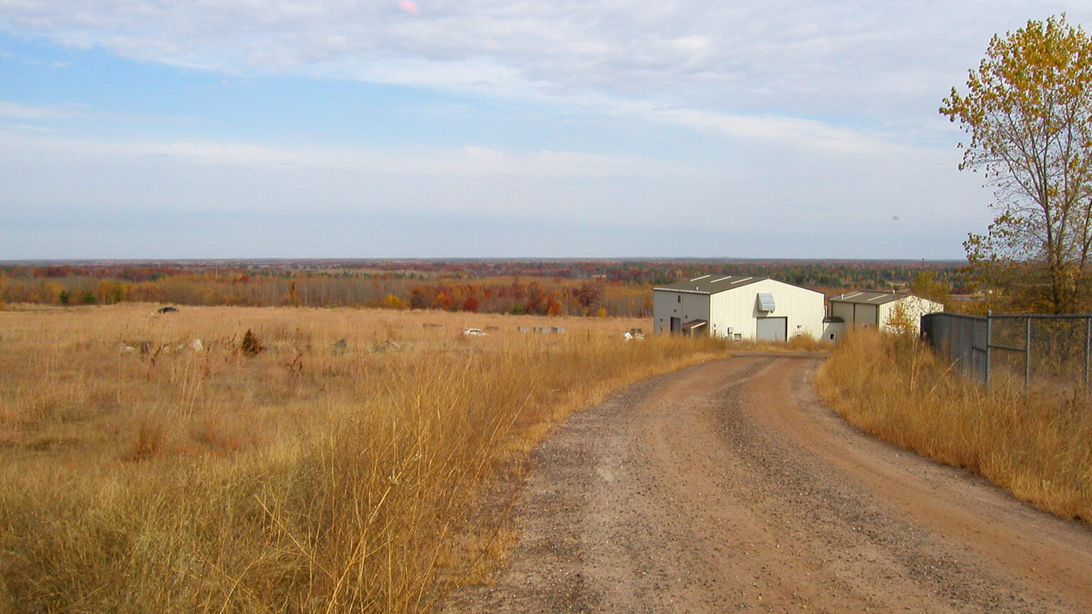 Two small warehouse buildings stand at the end of a gravel road, with a field in the foreground and trees with autumn leaves in the background extending to a distant horizon.
