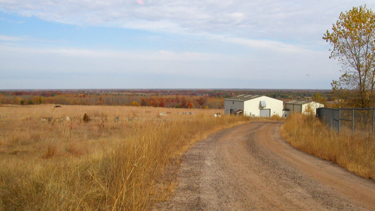 Two small warehouse buildings stand at the end of a gravel road, with a field in the foreground and trees with autumn leaves in the background extending to a distant horizon.