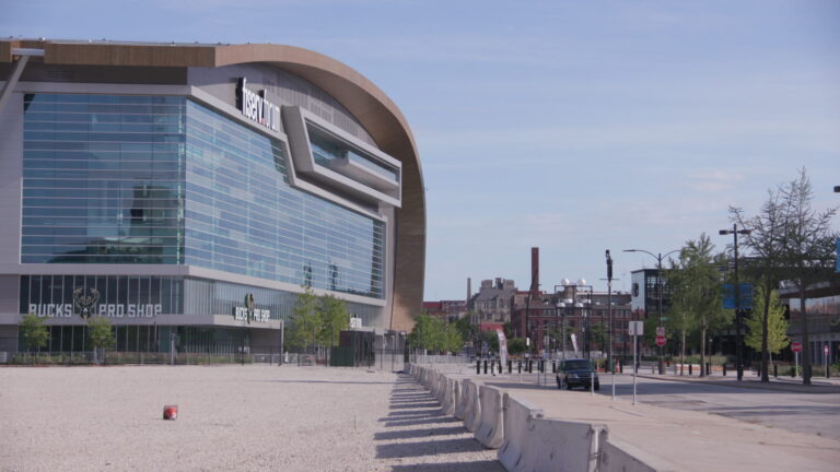 The glass facade of the Fiserv Forum overlooks an empty streetscape with buildings and trees in the background.