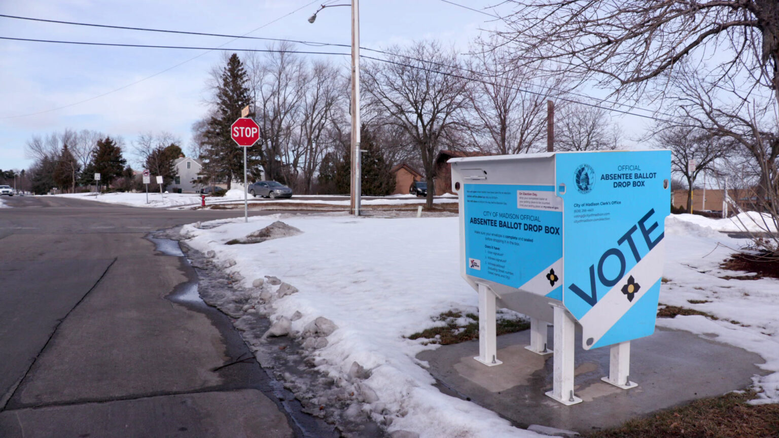 An absentee ballot drop box bolted to concrete next to an intersection stands amid melting snowdrifts, with trees and buildings in the background.