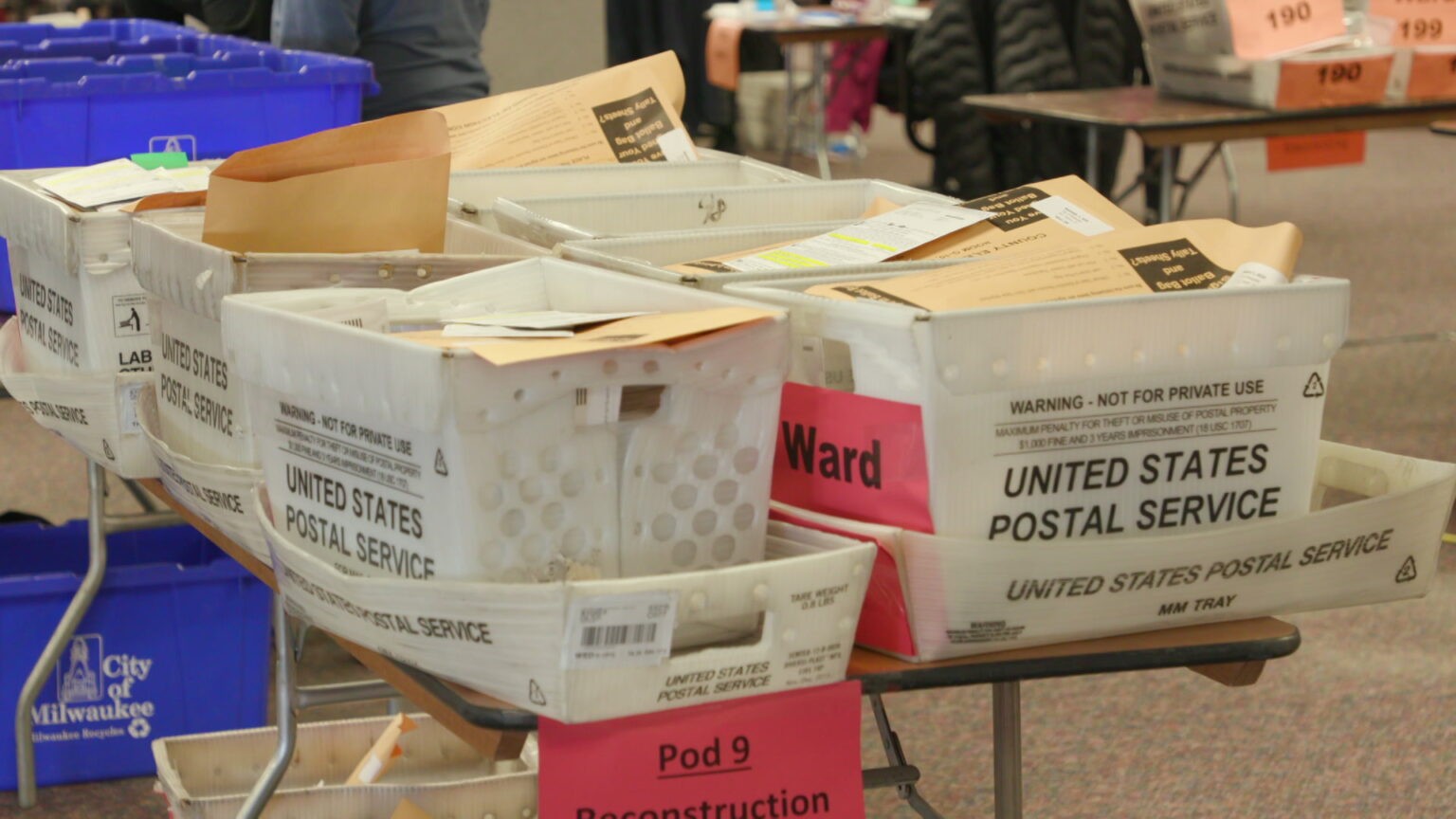 United States Postal Service boxes filled with absentee ballots sit on a table in a room.