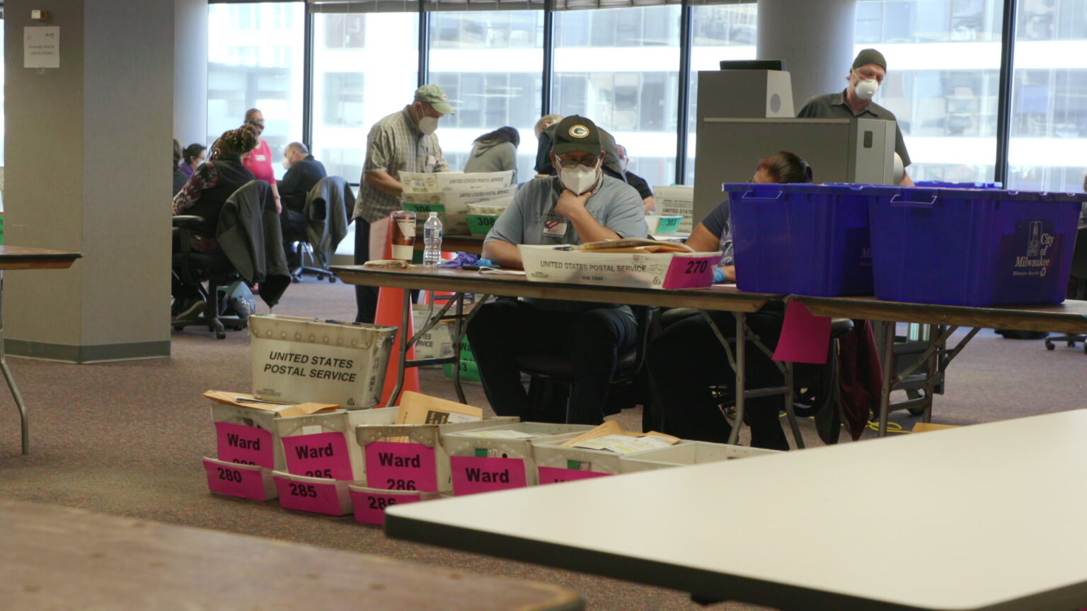 Election workers sit and stand at tables in a room with United States Postal Service boxes filled with absentee ballots sitting on the floor in the foreground.