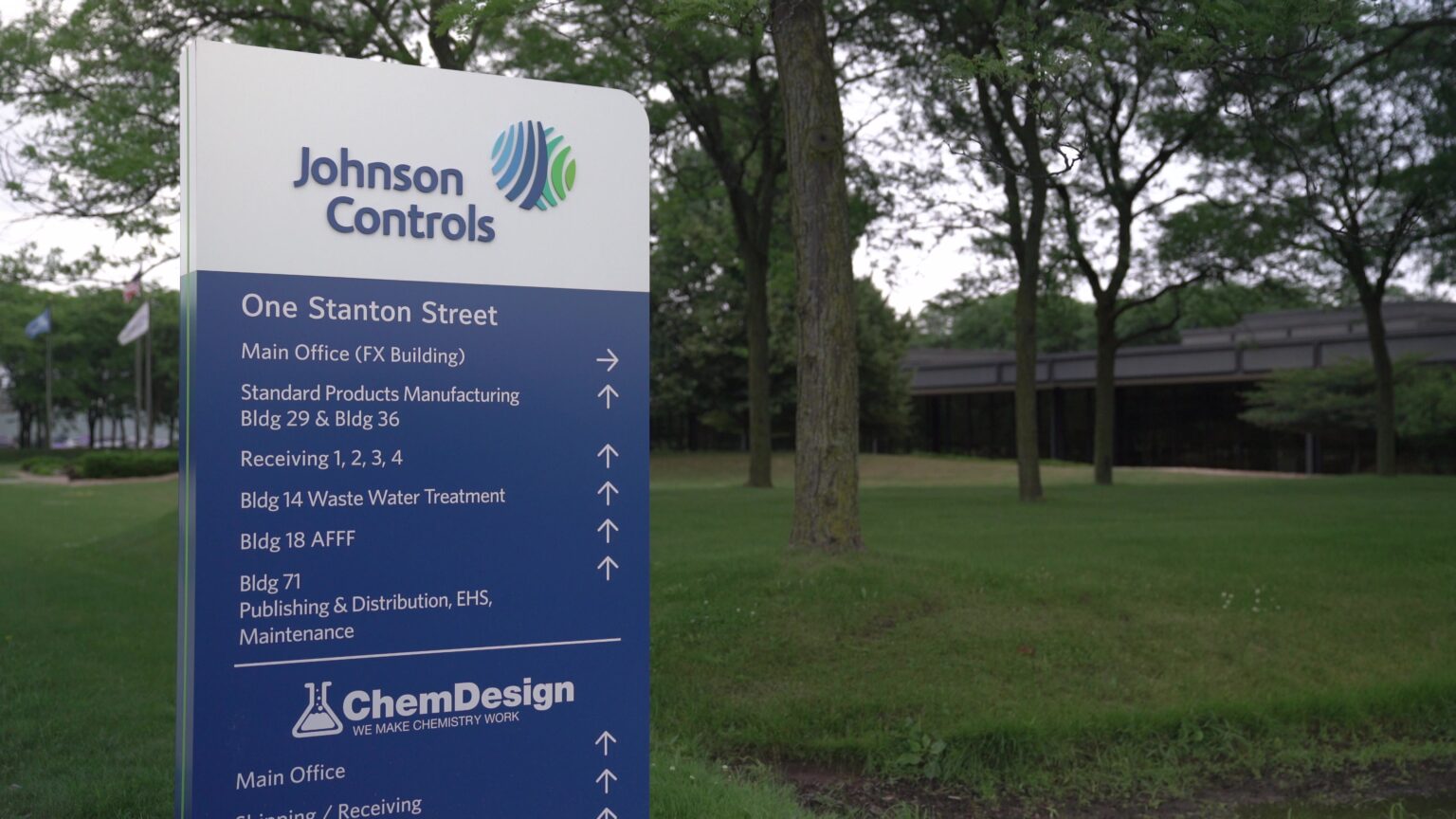 A sign with the Johnson Controls logo offers directions to different buildings, with trees, flagpoles and a low structure in the background.