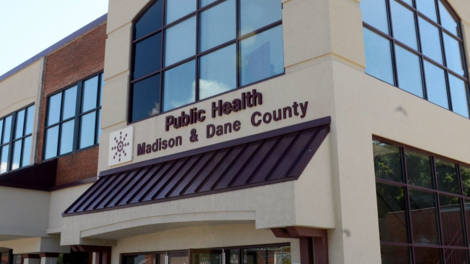 A two-story building with a concrete, brick and glass exterior is fronted with a sign that reads Public Health Madison & Dane County.