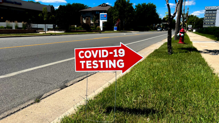 A yard sign in the shape of an arrow and with the words COVID-19 testing stands in a strip of grass between a street and sidewalk, with buildings, trees and business signs in the background.