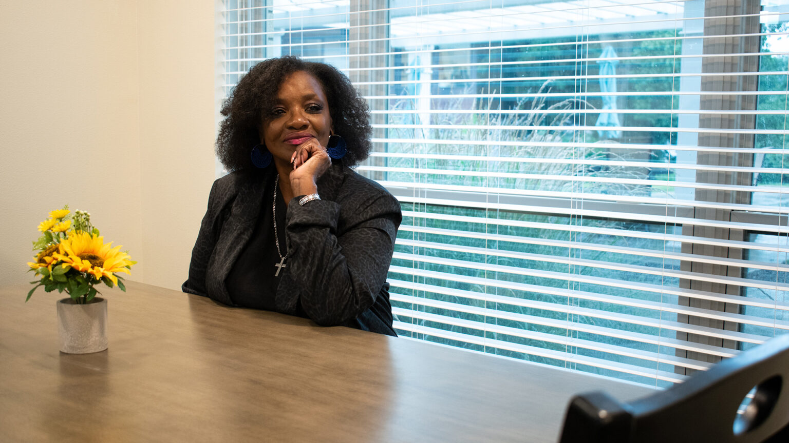 Kathann Jackson sits at a table with a small vase of flowers and a window with open blinds behind her.