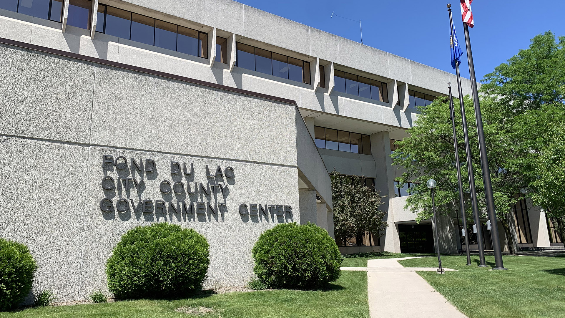 Flagpoles flying the U.S. and Wisconsin flags stand near trees and bushes in front of a multi-story concrete building with a sign reading "Fond du Lac City County Government Center."