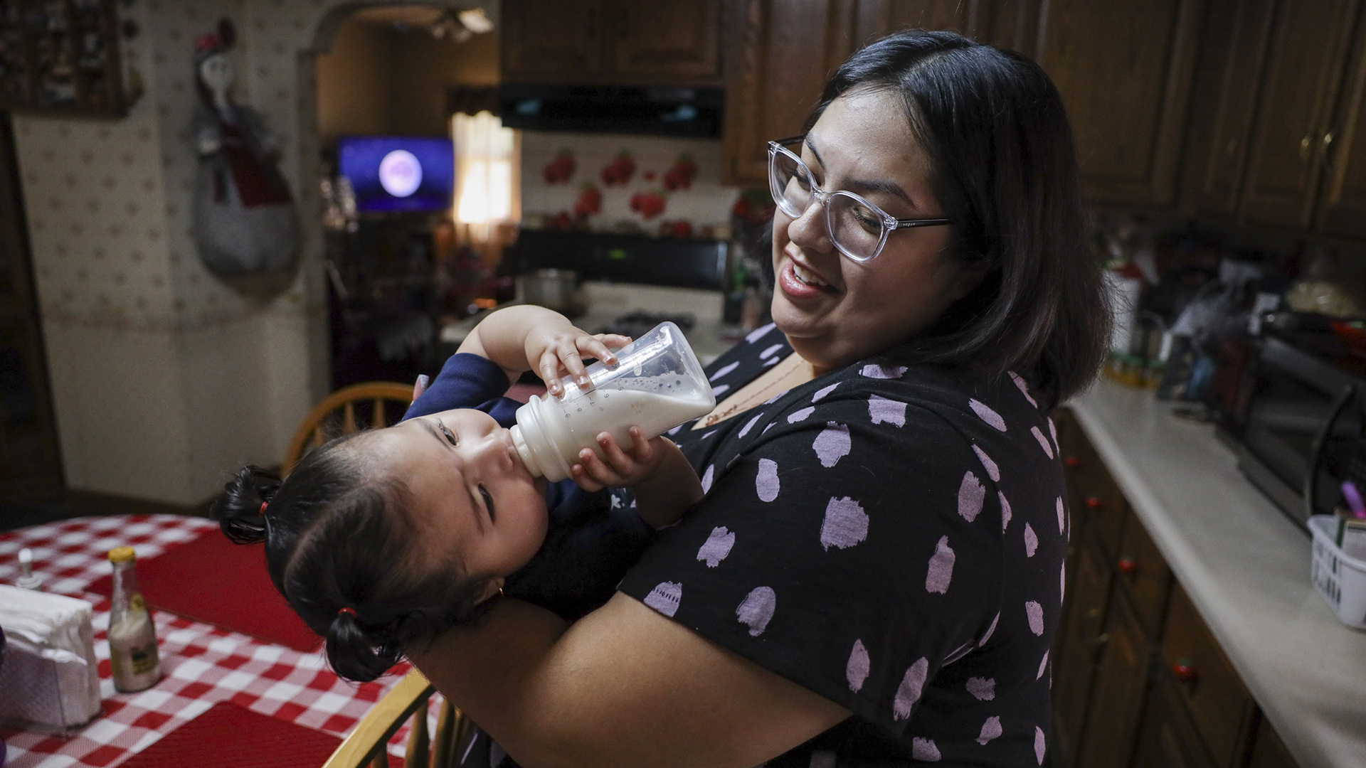 Adaliz Angeles feeds from a bottle while being held by Raquel Urbina while standing in a kitchen.