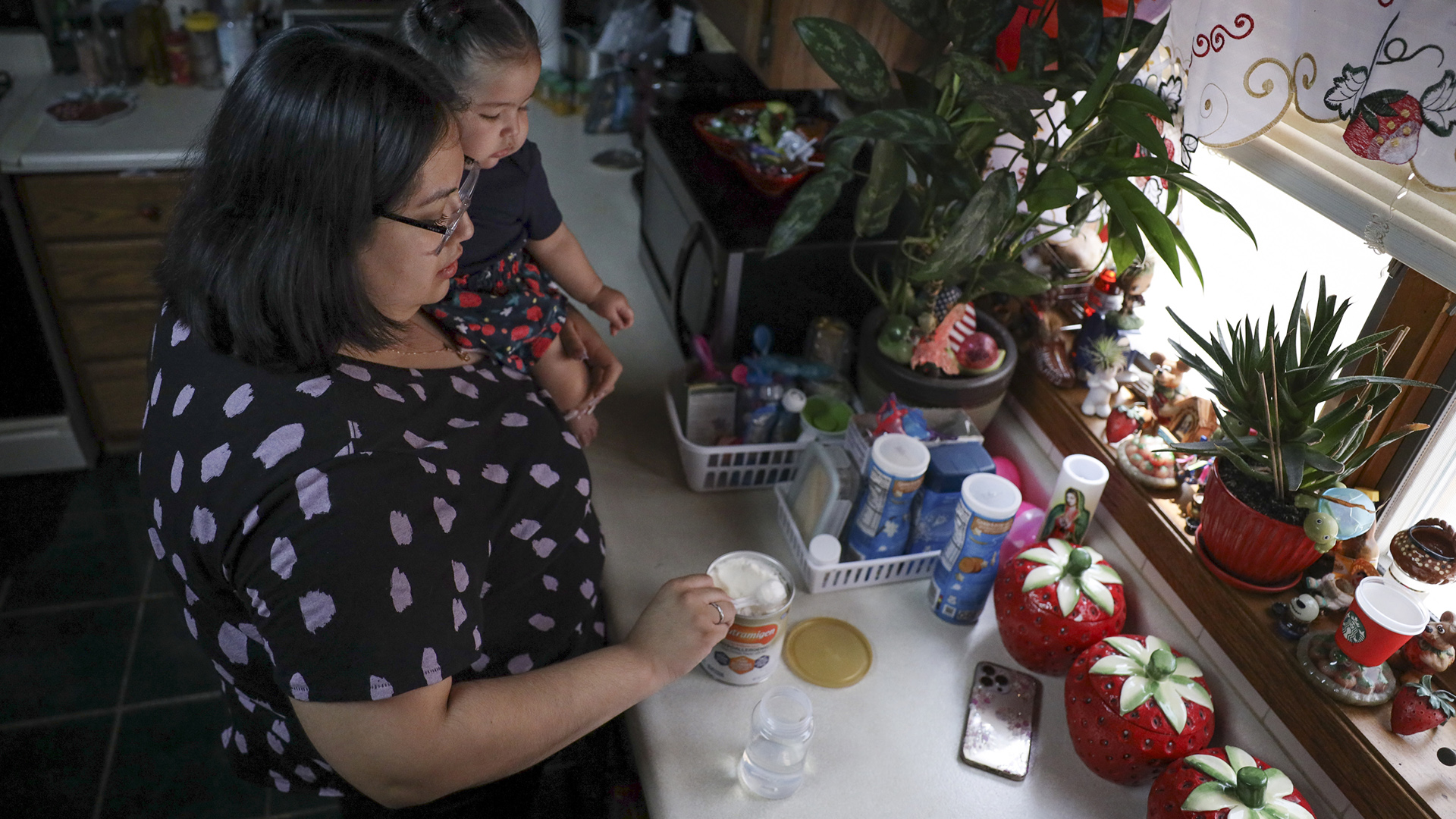 Raquel Urbina holds Adaliz Angeles in her left arm while preparing a bottle of formula on a kitchen counter.