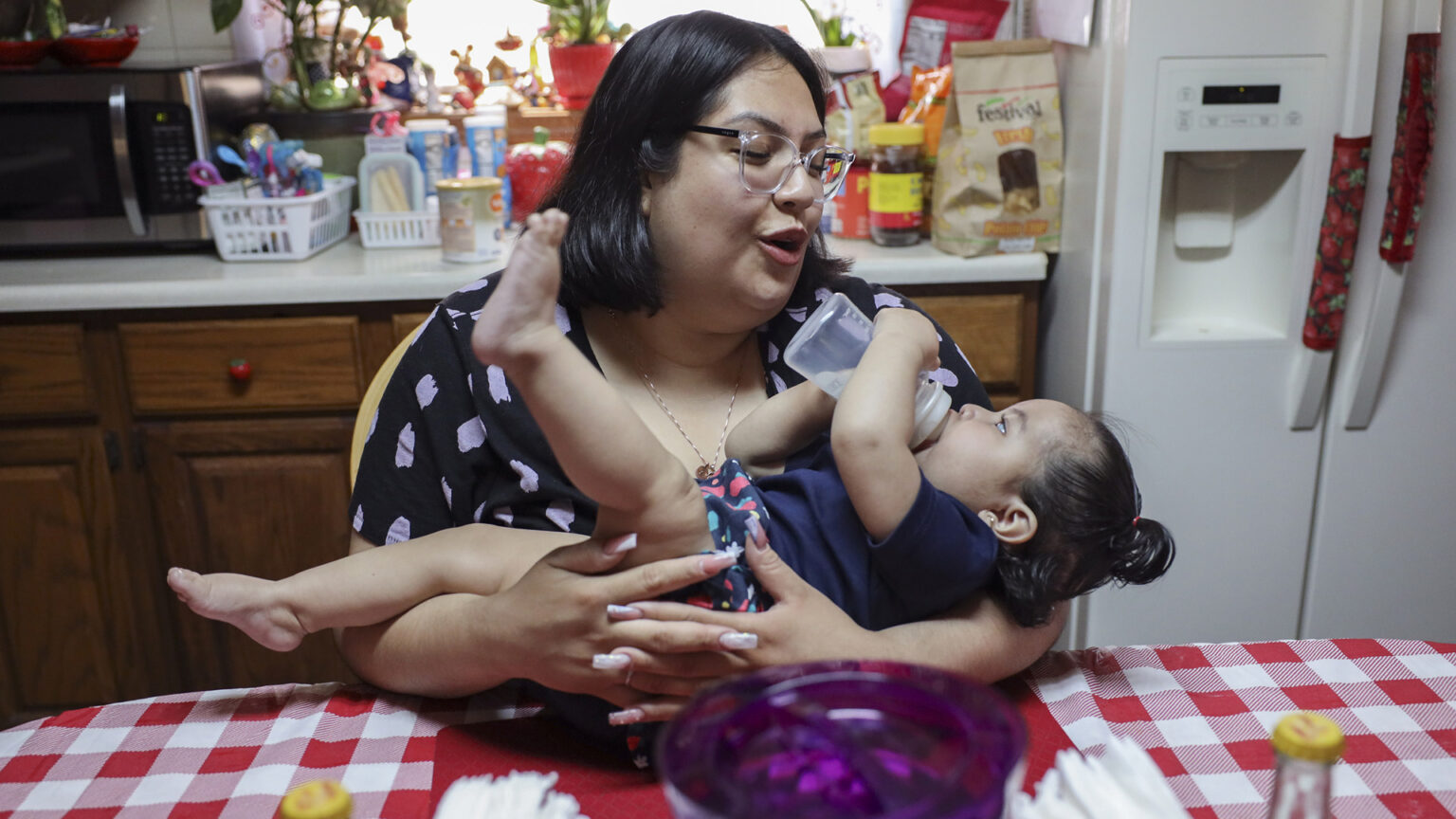 Raquel Urbina holds Adaliz Angeles at a kitchen table as the infant feeds on formula from a bottle, with a kitchen counter, cabinets and refrigerator in the background.