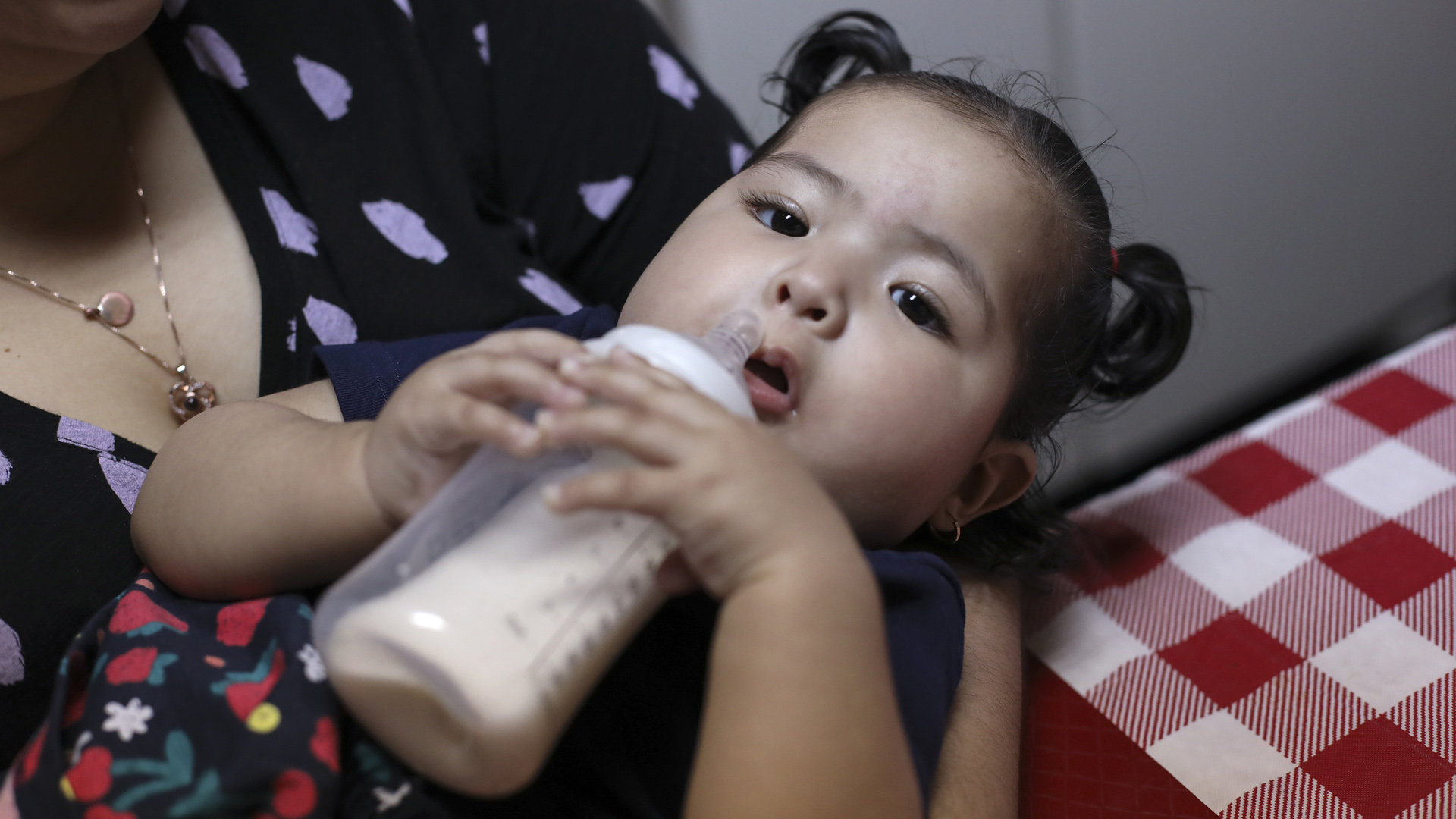 Adaliz Angeles holds a bottle to her mouth while being held at the edge of a table.