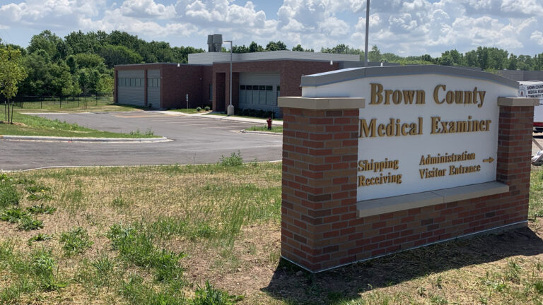 Brick columns frame a sign reading Brown County Medical Examiner that stands in front of a driveway to a brick building with high windows.