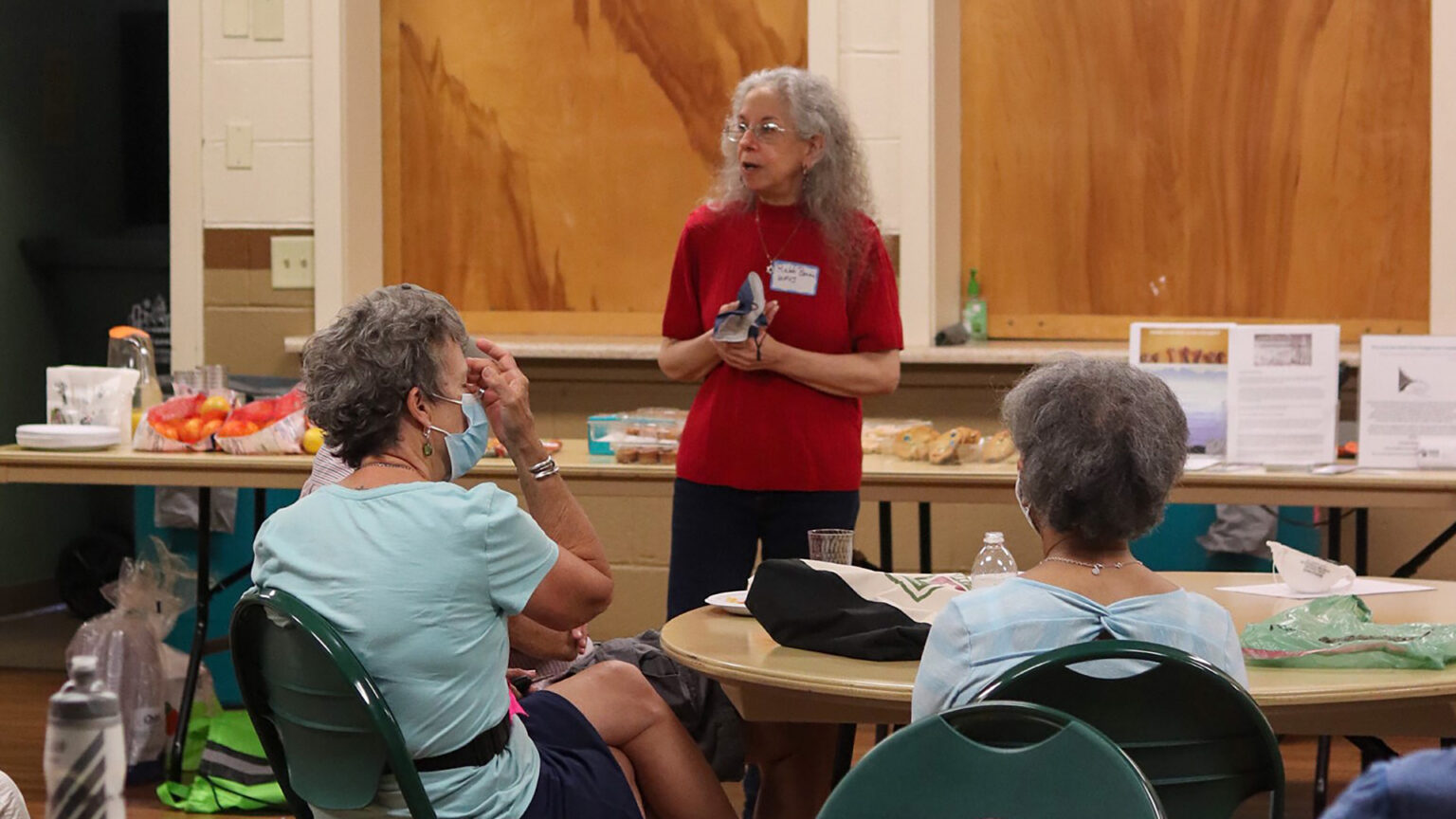 Bonnie Margulis stands in front of a table covered with food and other items and speaks to people seated at tables in a room with painted concrete block walls and wood-paneled cafeteria window coverings.