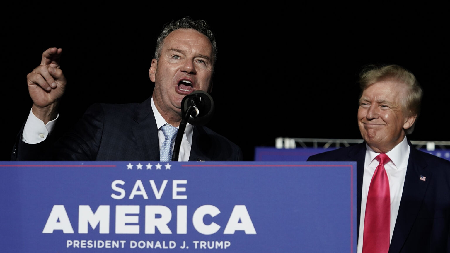 Tim Michels points with the index finger of his right hand while speaking into a microphone behind a podium printed with the words Save America and President Donald J. Trump while the Donald Trump stands to his left under a night sky.