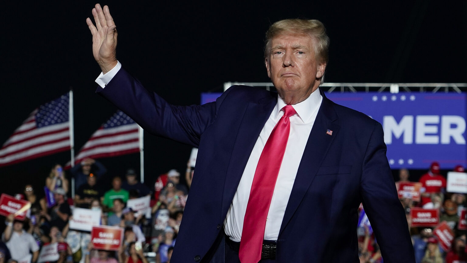 Donald Trump waves with his right hand while people sit on risers under a sign and U.S. flags in the background.