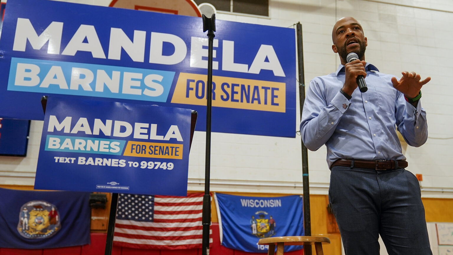 Mandela Barnes stands and speaks into a wireless microphone in a gymnasium, with signs reading Mandela Barnes for Senate and the U.S. and Wisconsin flags in the background.