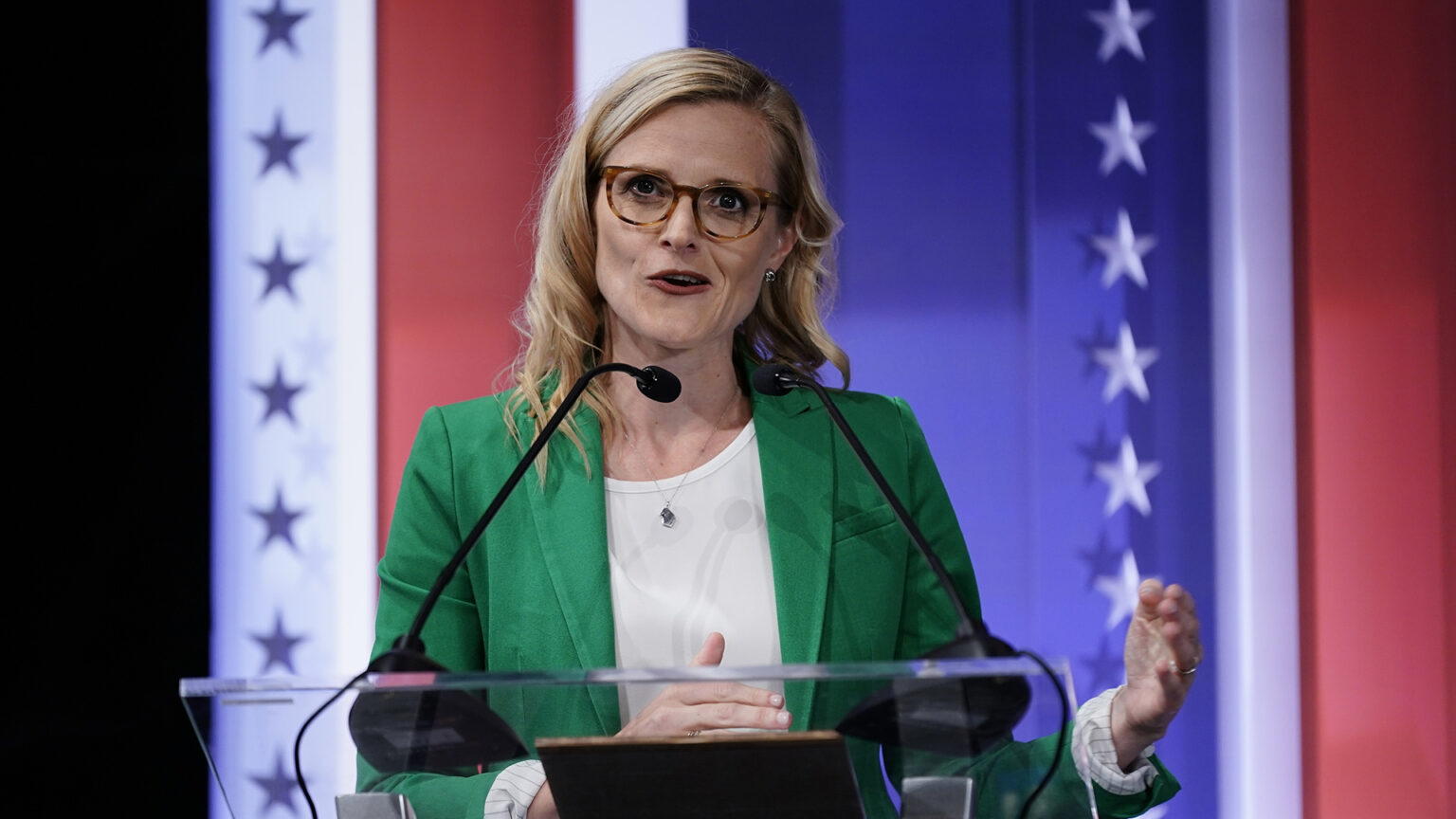 Sarah Godlewski speaks into a microphone while gesturing with both hands just below shoulder height with a stars-and-stripes-themed graphic in the background.
