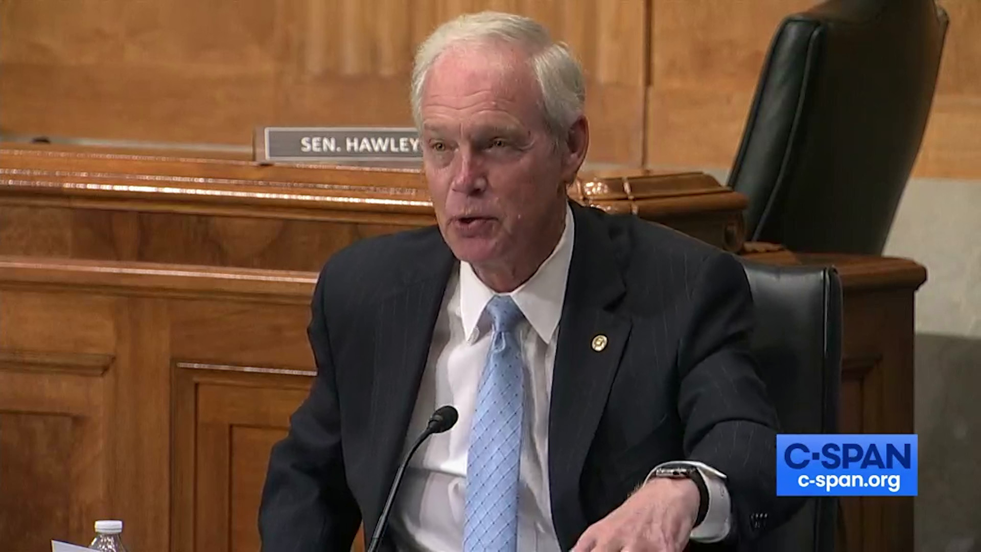 Ron Johnson speaks into a microphone in a legislative hearing room with an empty chair behind him.