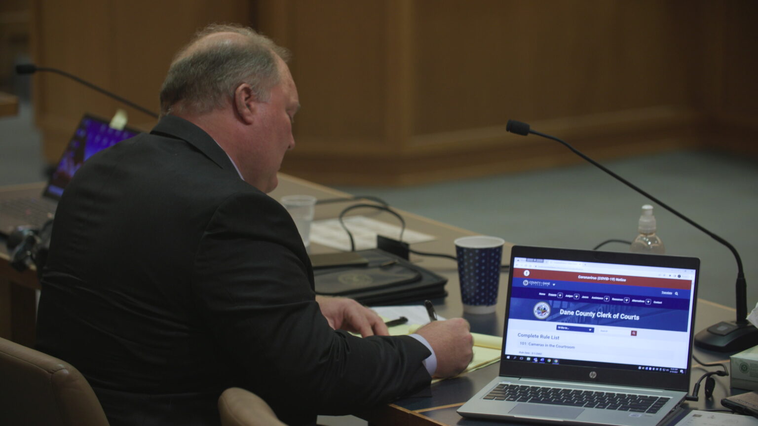 Michael Gableman sits at a counsel table in a courtroom and writes on a legal pad amid open laptops, coffee and water cups, and other items.