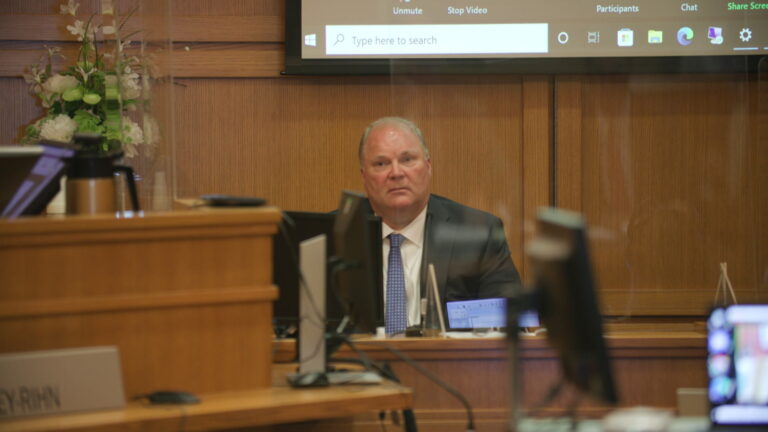 Michael Gableman sits in a witness box in a courtroom with the judge's bench to his right and the bottom of a large screen showing virtual participants in the background.