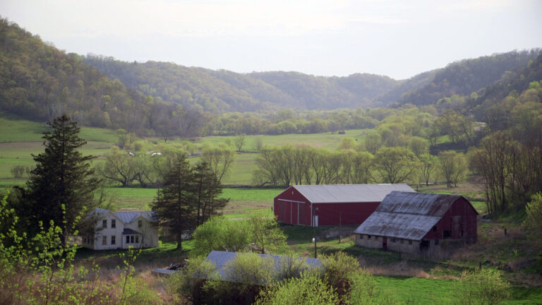 A farmhouse, barns and other outbuildings stand amid trees and fields in a valley, with low, wooded hills in the background.