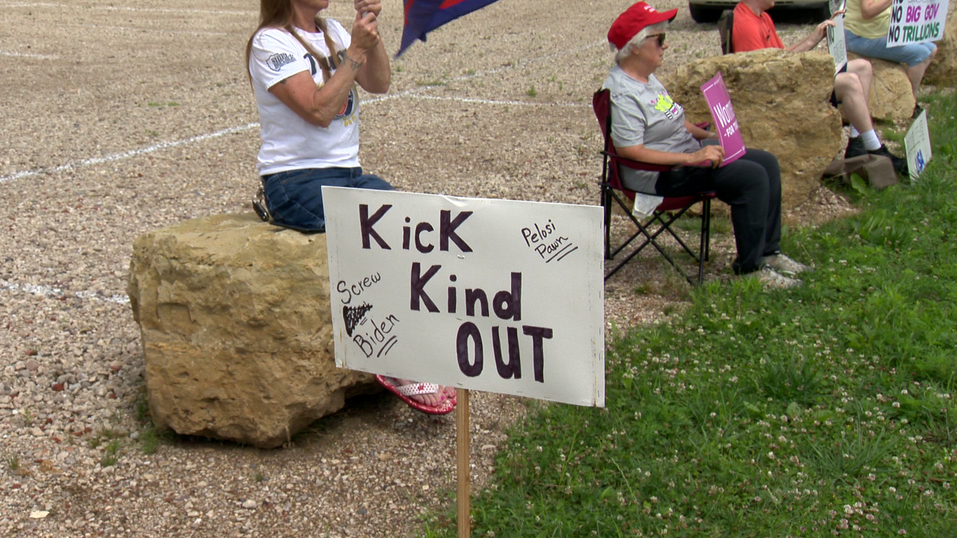 People sitting on boulders and folding lawn chairs hold signs in protest of Joe Biden and Ron Kind, with a sign planted in the grass in the foreground reading "Kick Kind Out," "Screw Biden" and "Pelosi Pawn."
