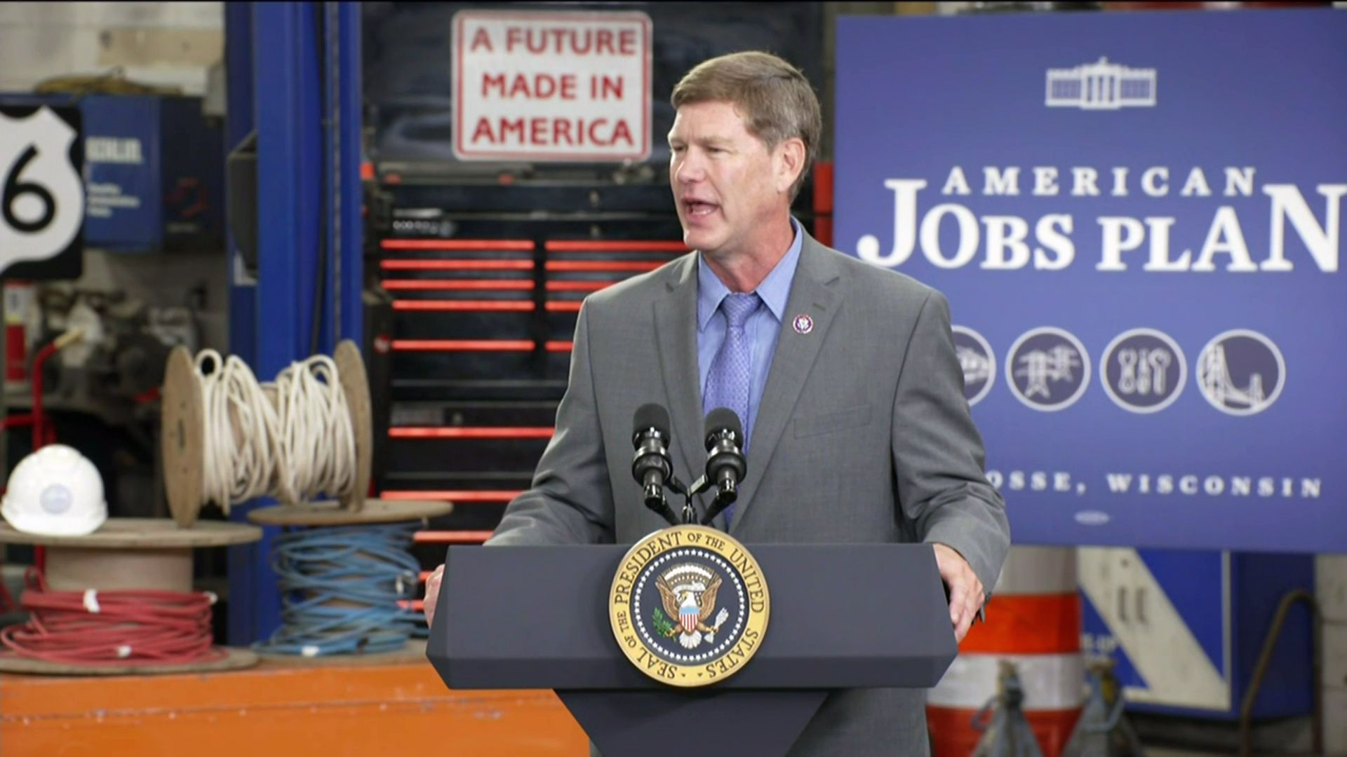 Ron Kind stands behind a podium with two microphones and the seal of the president of the United States inside a workplace with a tool cabinet, spools of electrical wire and other equipment, with signs in the background reading "American Jobs Plan" and "A Future Made in America."