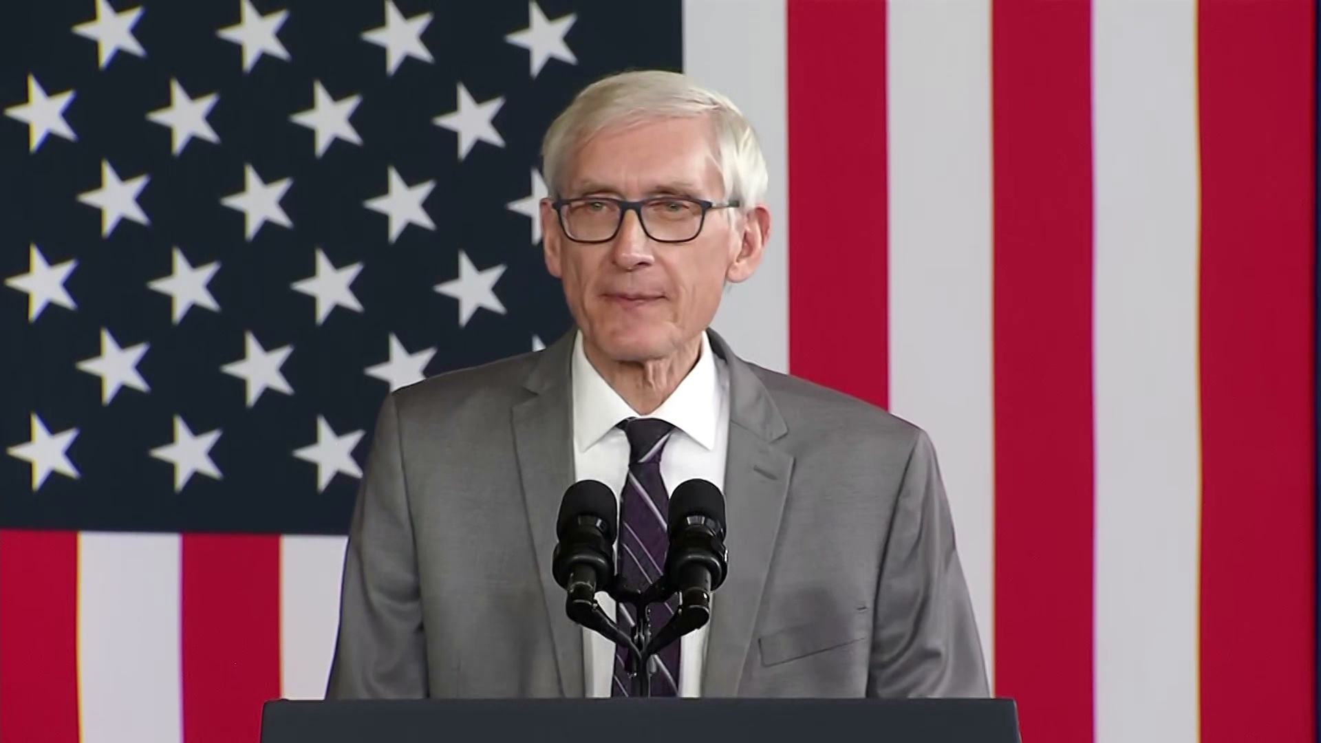 Tony Evers stands in front of a podium with two microphones in front of the backdrop of a U.S. flag.