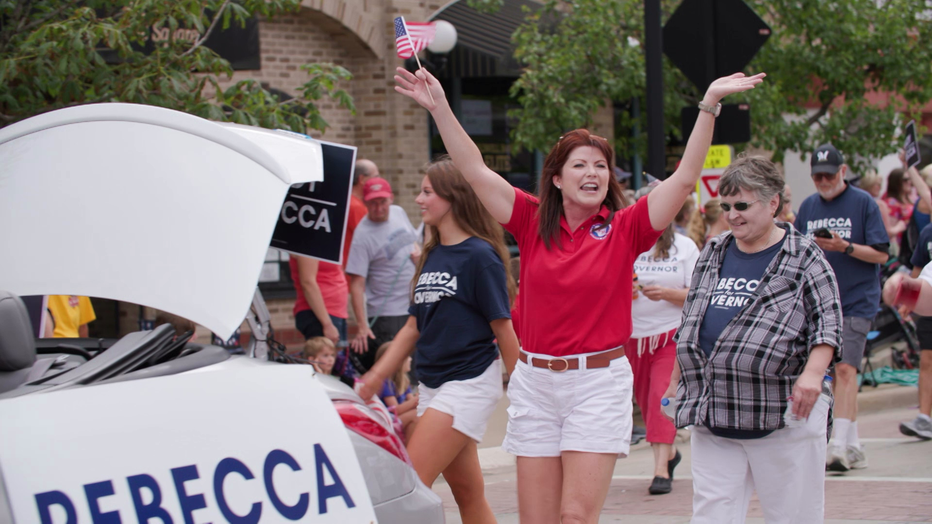 Rebecca Kleefisch holds her arms up in the air while holding a small U.S. flag in her right hand while walking down the street as part of a parade, surrounded by other people wearing t-shirts reading "Rebecca for Governor."