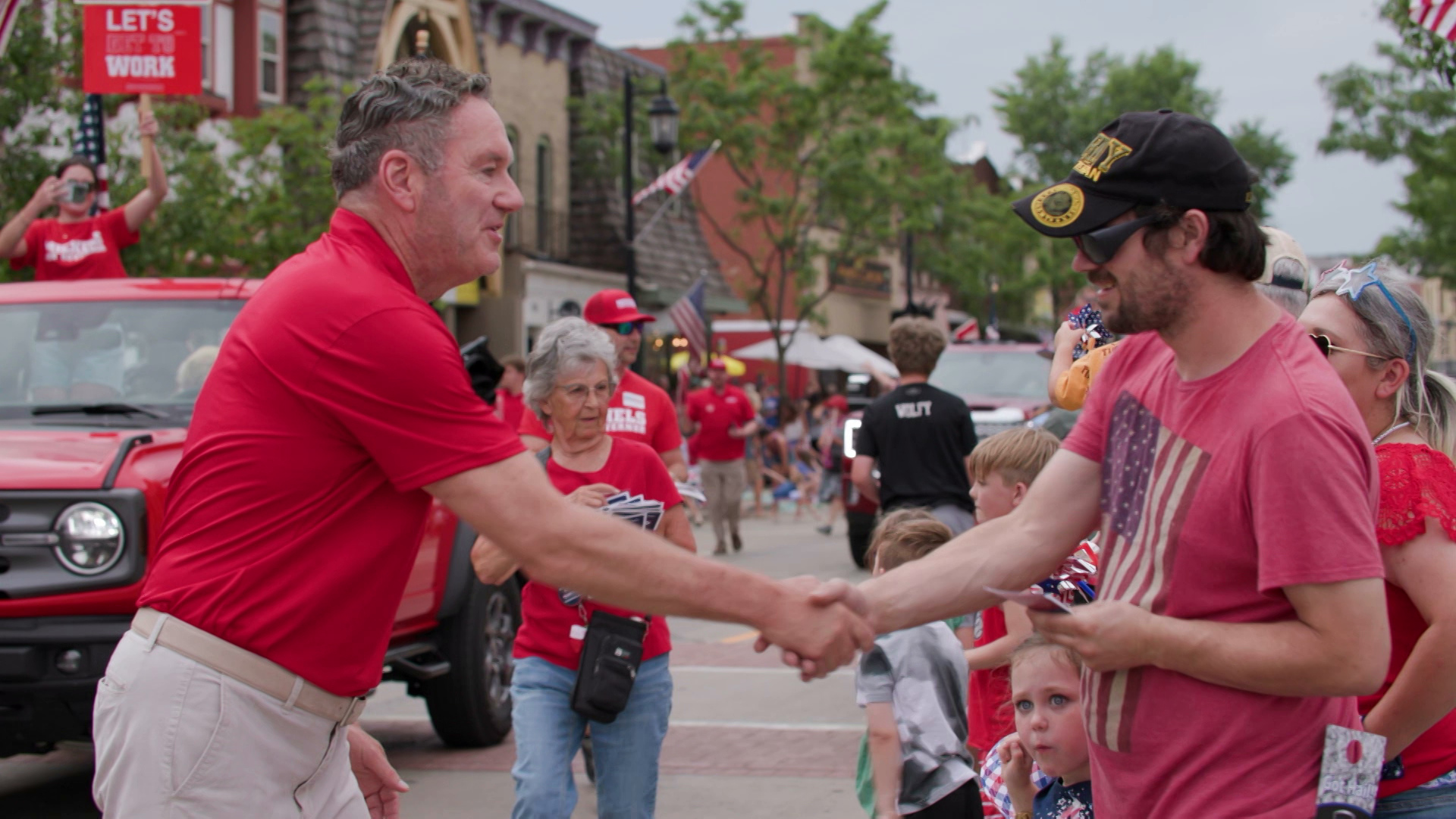 Tim Michels shakes the hand of another person while walking down a street as part of a parade.