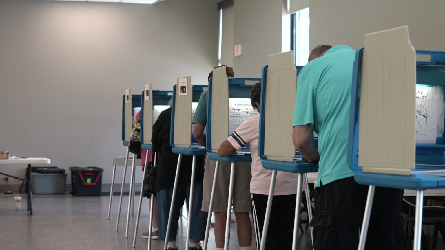 Voters fill out their ballots while standing in a row of privacy cubicles in a large room with blank walls.